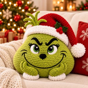 May include: A lime green crocheted pillow shaped like a character with a grumpy expression, large green eyes, and a black nose. It wears a red and white crocheted Santa hat with a white pom-pom and a small green holly leaf accent with red berries.