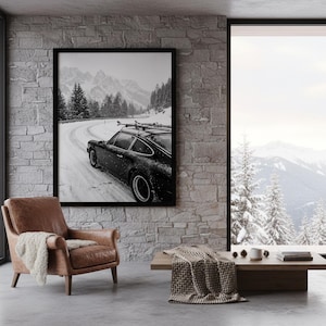 May include: A black and white framed print of a sports car driving on a snowy mountain road. The car has skis on the roof. The room has a leather armchair, a wooden coffee table, and a large window with a mountain view.