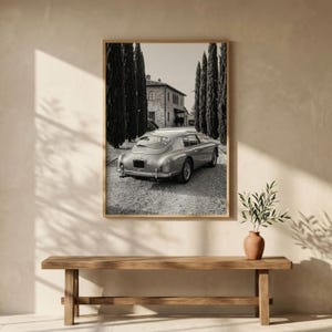 May include: A black and white framed wall art print depicting a vintage sports car parked on a gravel driveway in front of a stone building, flanked by tall, slender cypress trees. The scene captures the rear view of the car in a landscape.