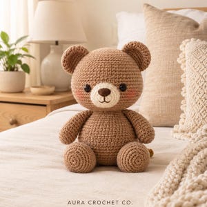 Può includere: Un orsacchiotto lavorato all'uncinetto di colore marrone chiaro con muso color crema, naso marrone scuro, occhi neri e guance rosate. L'orso è seduto e presenta la scritta AURA CROCHET CO. con un piccolo cuore rosa stampato sotto.