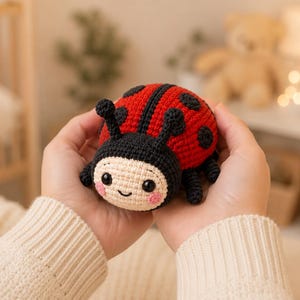 Puede incluir: Un peluche de mariquita tejido a ganchillo con el dorso rojo brillante y puntos negros, cabeza negra con dos antenas y cara de color crema con ojos negros y mejillas rosadas. El pequeño insecto tiene un cuerpo redondeado y patas negras.