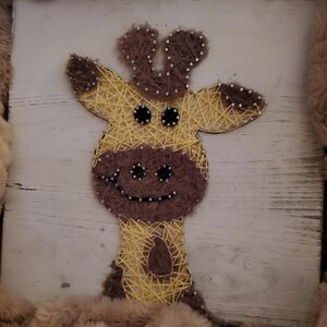 May include: A string art giraffe on a white wooden background. The giraffe is made with yellow and brown thread, with brown accents for the ears, nose, and spots. The giraffe has a smiling face and is framed by a fluffy, brown border.
