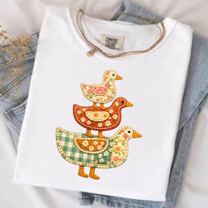 May include: White t-shirt featuring a stack of three decorative ducks. The ducks have floral and plaid patterns in shades of orange, green, and pink. The shirt has a Comfort Colors tag.