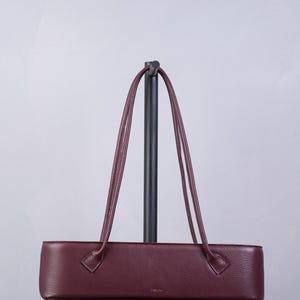 May include: A long, rectangular, burgundy-colored handbag with a textured surface and two thin, matching shoulder straps. The bag features a small, gold-toned logo centered on the front panel and is displayed on a black vertical stand.