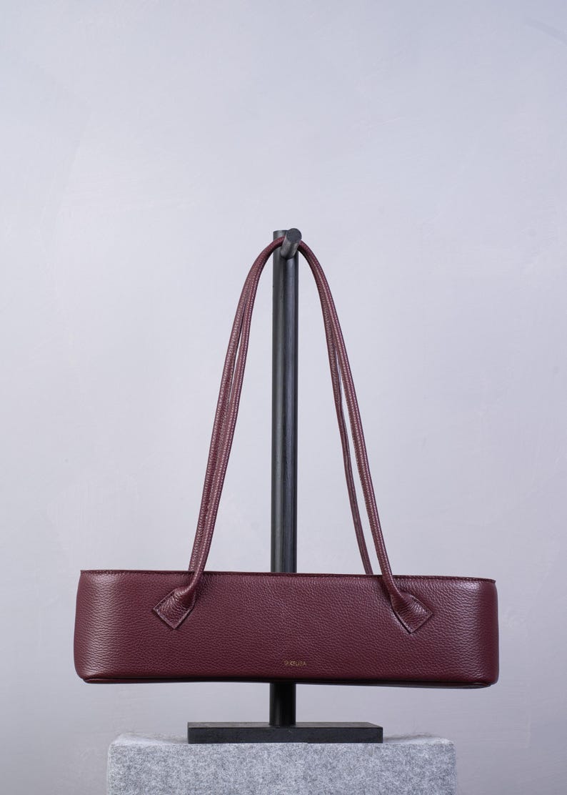 May include: A long, rectangular, burgundy-colored handbag with a textured surface and two thin, matching shoulder straps. The bag features a small, gold-toned logo centered on the front panel and is displayed on a black vertical stand.