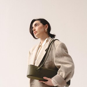 May include: A structured olive green shoulder bag with a textured finish and a long strap, worn over a beige tweed blazer with a white collared shirt underneath. The bag features a rectangular shape with rounded edges and a minimalist design.