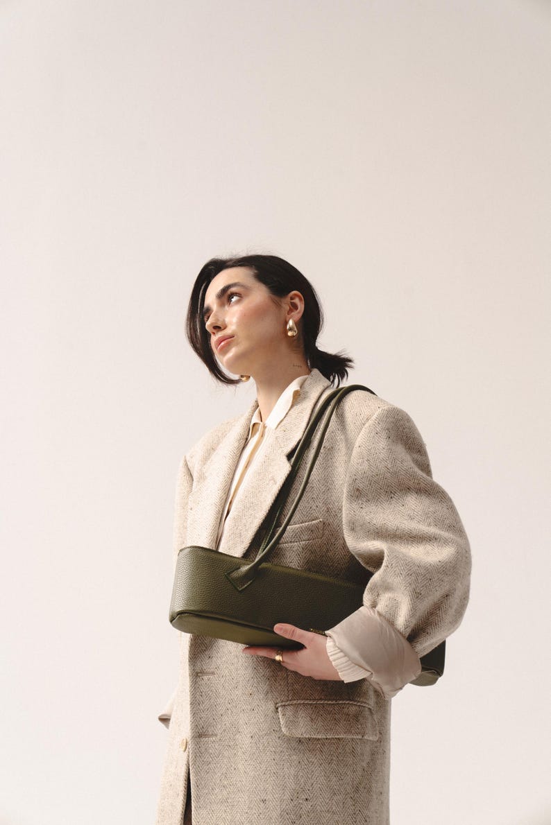 May include: A structured olive green shoulder bag with a textured finish and a long strap, worn over a beige tweed blazer with a white collared shirt underneath. The bag features a rectangular shape with rounded edges and a minimalist design.