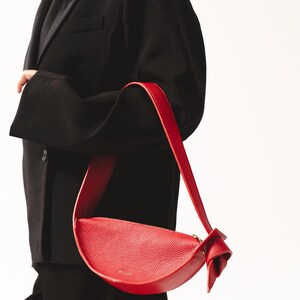 May include: A bright red crescent-shaped shoulder bag with a textured pebbled surface, a single wide strap, and a knotted detail on one side. The bag features a top zipper closure and gold-toned hardware, presenting a curved silhouette.