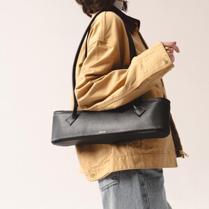 May include: A black, horizontally elongated, rectangular shoulder bag with a pebbled leather texture. The bag features a top zipper closure, two thin black shoulder straps, and the gold-toned text "YAGUBA" printed on the front center.