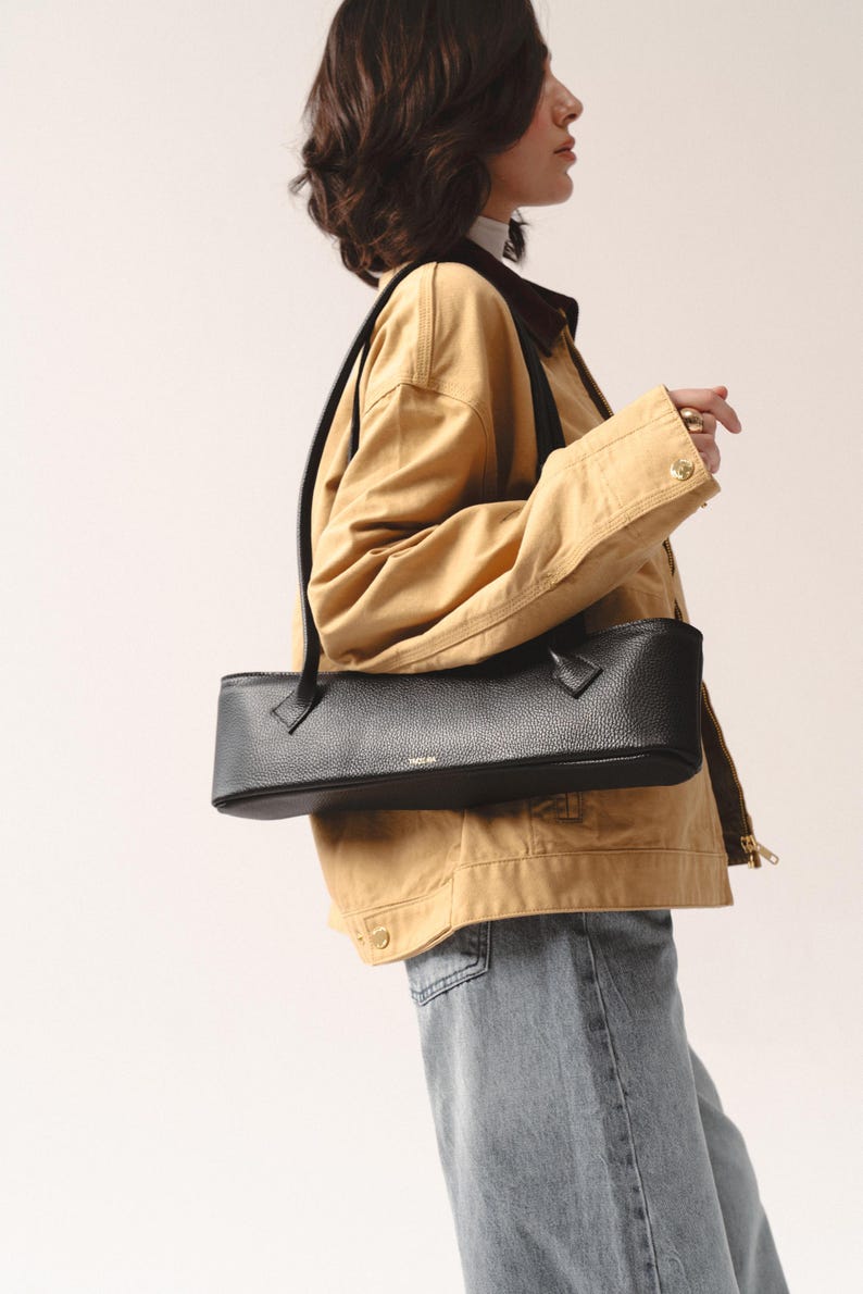 May include: A black, horizontally elongated, rectangular shoulder bag with a pebbled leather texture. The bag features a top zipper closure, two thin black shoulder straps, and the gold-toned text "YAGUBA" printed on the front center.