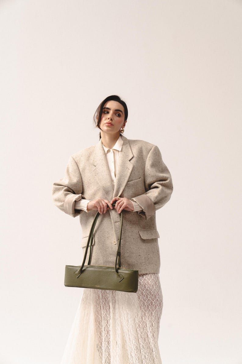 May include: An olive green rectangular shoulder bag with thin straps, worn with an oversized beige speckled blazer, a white collared shirt, and a white lace skirt. The bag features a structured, elongated horizontal shape and a smooth finish.