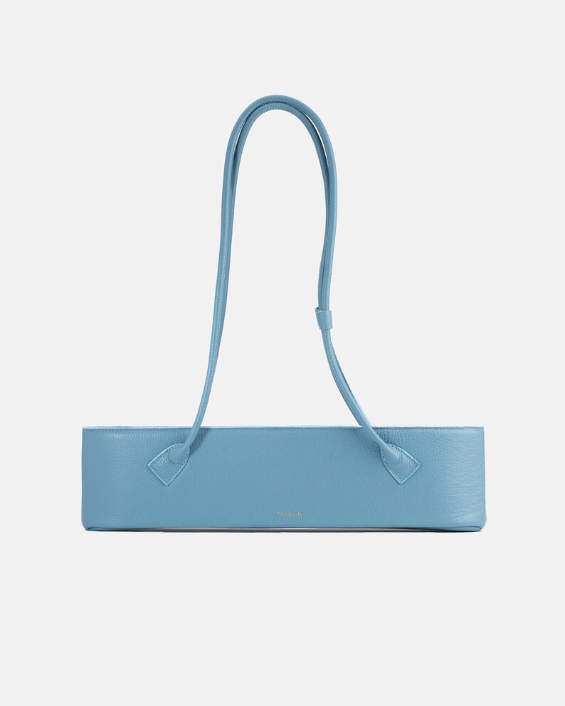May include: A light blue, horizontally elongated rectangular handbag with a textured surface. It features a single, long, thin shoulder strap attached to the top corners of the bag. The design is minimalist with a small, centered logo on the front.