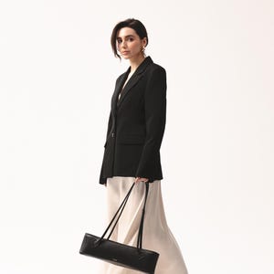 May include: A black blazer with a single button closure, a cream-colored midi skirt with a fluid drape, and black strappy high-heeled sandals. A long, rectangular black shoulder bag with thin straps is held at the side of the garment ensemble.