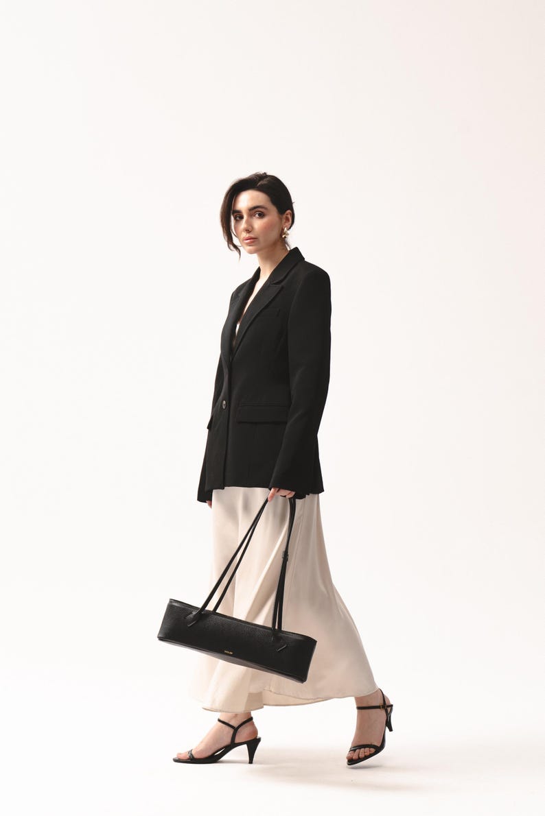 May include: A black blazer with a single button closure, a cream-colored midi skirt with a fluid drape, and black strappy high-heeled sandals. A long, rectangular black shoulder bag with thin straps is held at the side of the garment ensemble.