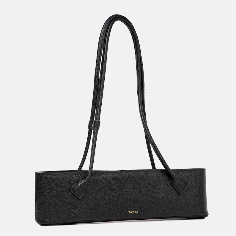 May include: A black, horizontally elongated shoulder bag with a pebbled leather texture. The bag features two long, thin, matching black shoulder straps and the brand name TAKSUBA printed in small gold lettering on the front lower center.