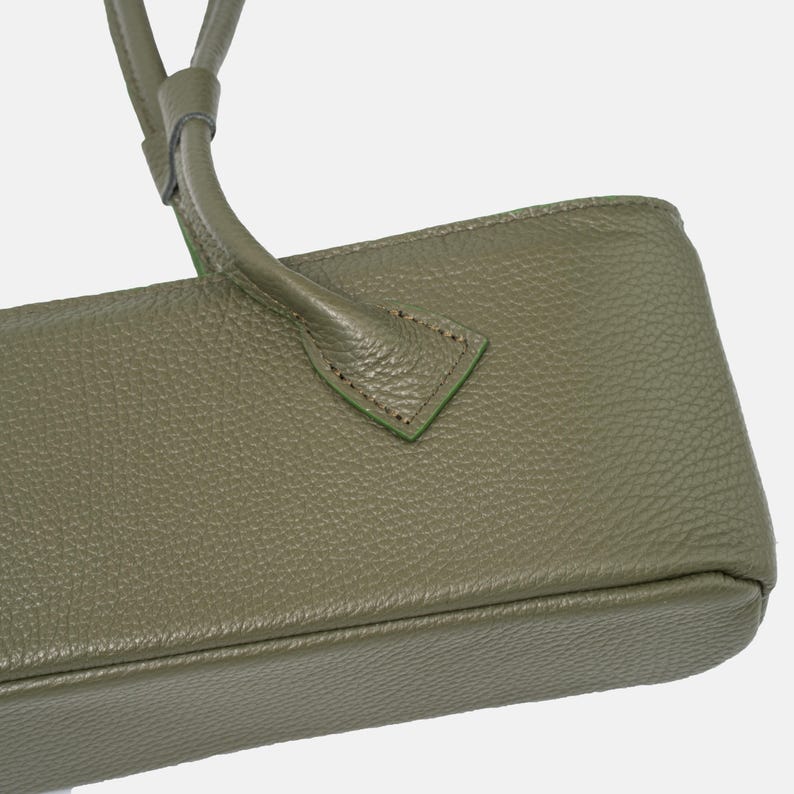 May include: An olive green handbag featuring a textured, pebbled surface. The bag includes a sturdy handle attached with triangular stitching details. The design showcases a clean, rectangular base structure with visible seams along the edges.