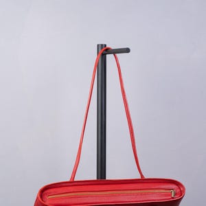 May include: A bright red leather handbag featuring a textured surface, a horizontal gold-toned metal zipper closure along the top edge, and two thin matching red leather shoulder straps attached to the sides of the rectangular bag body.