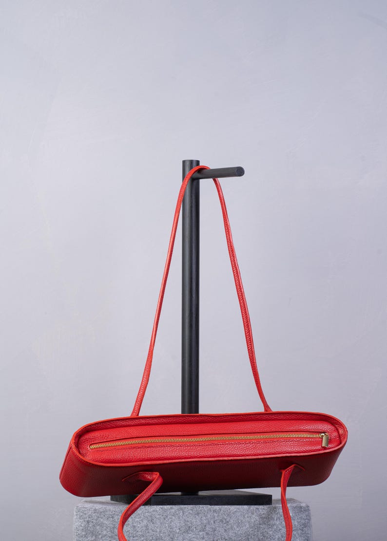 May include: A bright red leather handbag featuring a textured surface, a horizontal gold-toned metal zipper closure along the top edge, and two thin matching red leather shoulder straps attached to the sides of the rectangular bag body.