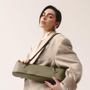 May include: An olive green, elongated, rectangular shoulder bag with a pebbled leather texture. The bag features two thin, matching shoulder straps and a small gold-toned logo embossed on the front center. The design is structured and horizontal.