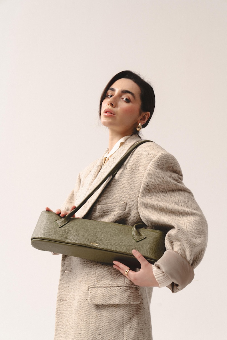 May include: An olive green, elongated, rectangular shoulder bag with a pebbled leather texture. The bag features two thin, matching shoulder straps and a small gold-toned logo embossed on the front center. The design is structured and horizontal.