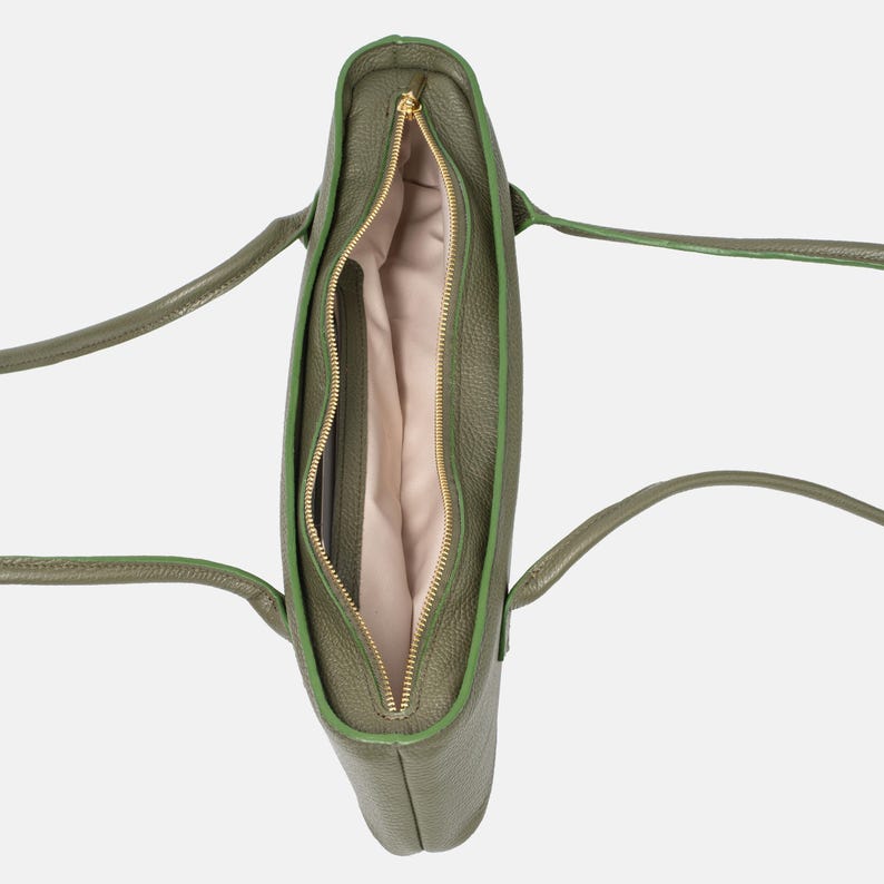 May include: An olive green tote bag with a textured exterior, featuring two long shoulder straps and a central gold-toned zipper closure. The interior is lined with a light beige fabric, revealing a single main compartment and a side pocket.