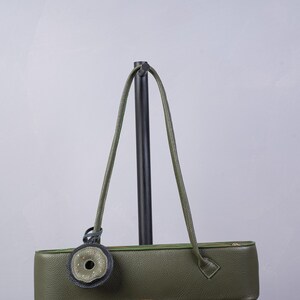 May include: An olive green leather shoulder bag with a long, rectangular, horizontal silhouette. The bag features a top zipper closure, two thin shoulder straps, and a circular decorative charm with a dark grey border and a central opening.