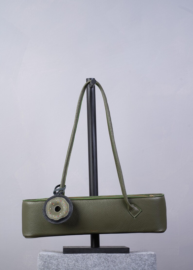May include: An olive green leather shoulder bag with a long, rectangular, horizontal silhouette. The bag features a top zipper closure, two thin shoulder straps, and a circular decorative charm with a dark grey border and a central opening.