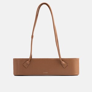 May include: A tan, rectangular, structured shoulder bag with a pebbled texture. The bag features two long, thin, matching tan shoulder straps attached to the front panel. The word "TAIZUZA" is printed in small, centered, silver-toned lettering.