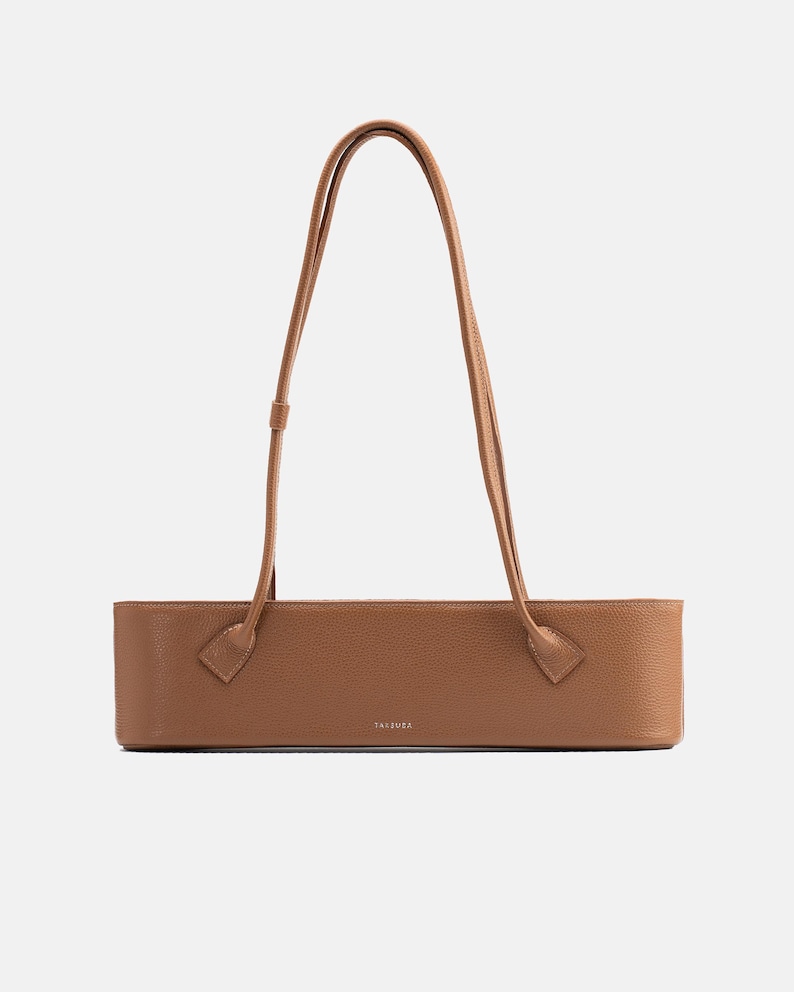 May include: A tan, rectangular, structured shoulder bag with a pebbled texture. The bag features two long, thin, matching tan shoulder straps attached to the front panel. The word "TAIZUZA" is printed in small, centered, silver-toned lettering.