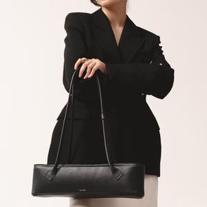 May include: A black, horizontally elongated, rectangular shoulder bag with a pebbled texture and two thin, long straps. The bag features a small, gold-toned logo printed on the front center. The design is structured with clean, straight lines.