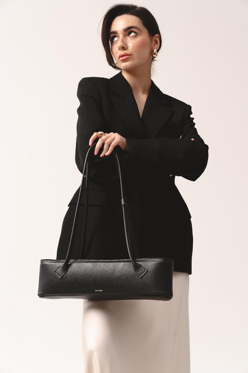 May include: A black, horizontally elongated, rectangular shoulder bag with a pebbled texture and two thin, long straps. The bag features a small, gold-toned logo printed on the front center. The design is structured with clean, straight lines.