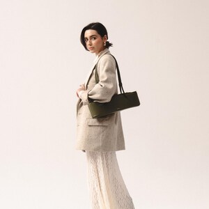 May include: A long, rectangular olive green shoulder bag with a slim profile and thin straps, worn over a beige speckled blazer and a long, white, textured lace skirt. The bag features a smooth finish and a small, centered gold-toned logo detail.