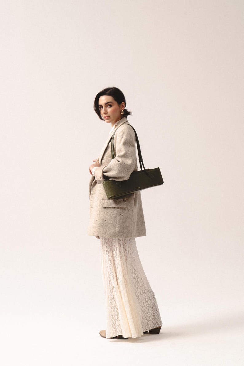 May include: A long, rectangular olive green shoulder bag with a slim profile and thin straps, worn over a beige speckled blazer and a long, white, textured lace skirt. The bag features a smooth finish and a small, centered gold-toned logo detail.