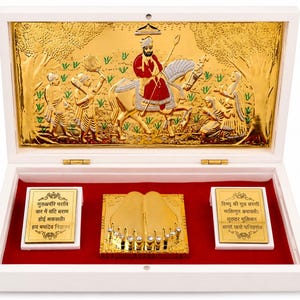 May include: A white wooden display box containing a gold-toned metal relief plaque of a figure on horseback, a central gold-toned metal pair of feet with embedded clear crystals, and two gold-toned metal plaques with black text in Hindi script.