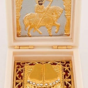May include: A cream-colored square box with a hinged lid. The interior lid features a gold and silver relief of a figure on horseback. The base contains a gold-toned plaque with a pair of feet adorned with small clear crystals on the toes.