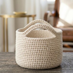 May include: A cream-colored, crocheted basket with two handles. The basket is cylindrical and sits on a wooden surface. The crochet stitches are visible, creating a textured appearance. The basket is empty.