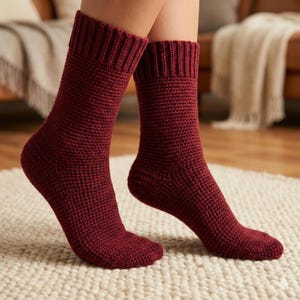 May include: A pair of burgundy-colored knitted socks. The socks have a ribbed cuff and a textured, waffle-like pattern throughout the body. The socks are shown on a light-colored rug.