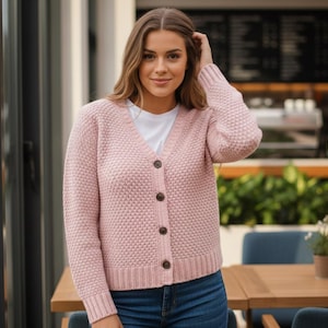 May include: A pale pink cardigan with a textured knit design and dark brown buttons. The cardigan is worn over a white t-shirt and paired with blue jeans. The woman is standing outside near a cafe.