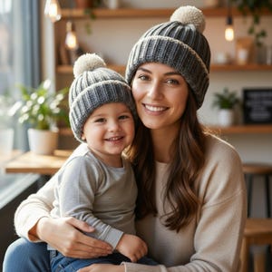Puede incluir: Una mujer y un niño con gorros de punto grises a juego con pompones blancos. La mujer lleva un suéter beige y el niño una camiseta gris de manga larga. Ambos sonríen.