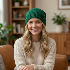 May include: A woman wearing a green knitted beanie and a cream-colored sweater. The beanie is a solid, vibrant green color and appears to be made of a chunky knit. The sweater has a ribbed texture. The background includes a wooden bookshelf and a leather couch.