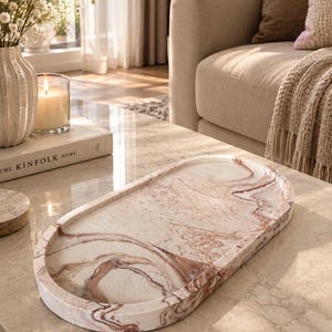 May include: An oval-shaped tray featuring a marbled pattern with swirls of cream, beige, and reddish-brown tones. The tray has a smooth, polished surface with a raised edge, showcasing a distinct stone-like appearance with organic color variations.