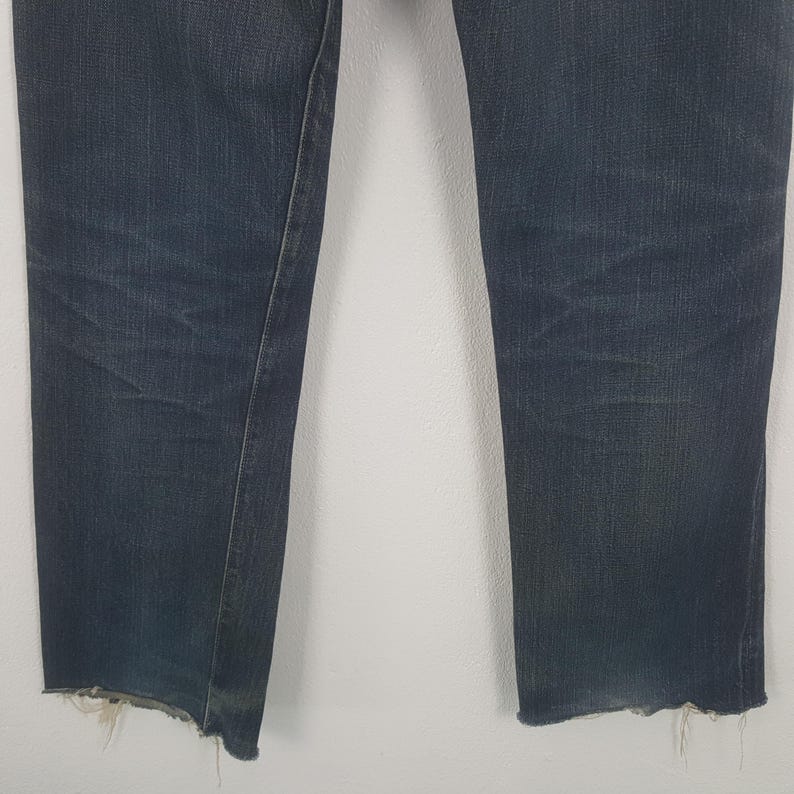 May include: Dark wash denim jeans with a raw, frayed hem. The jeans have a straight leg cut and show signs of wear. The fabric is a deep indigo blue with subtle fading. The jeans are a classic style.