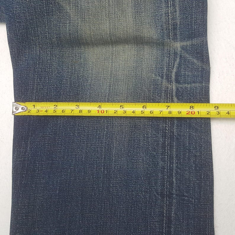 May include: Close-up of blue denim jeans with a yellow measuring tape. The tape shows measurements in inches. The jeans have a faded appearance and are on a white background. The tape measures from 1 to 9 inches.