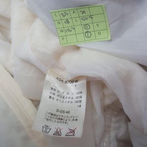 Pode incluir: Close-up de uma pe&ccedil;a de roupa branca com uma etiqueta. A etiqueta tem texto em japon&ecirc;s e ingl&ecirc;s, incluindo "ASK-9708 W" e detalhes sobre a composi&ccedil;&atilde;o do tecido. Uma etiqueta verde com n&uacute;meros manuscritos tamb&eacute;m &eacute; vis&iacute;vel.