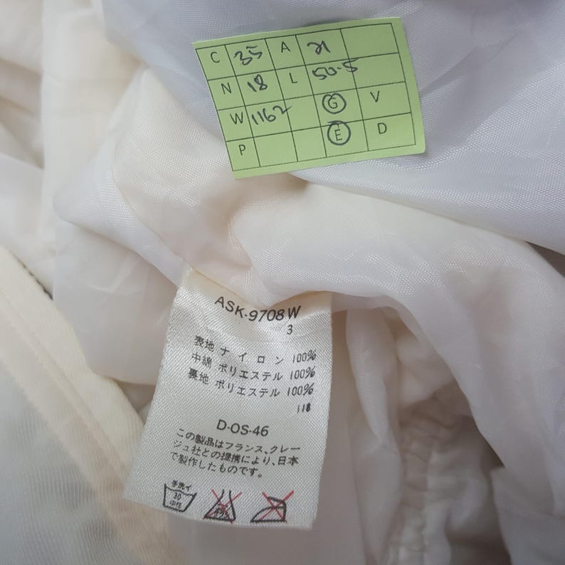 Pode incluir: Close-up de uma pe&ccedil;a de roupa branca com uma etiqueta. A etiqueta tem texto em japon&ecirc;s e ingl&ecirc;s, incluindo "ASK-9708 W" e detalhes sobre a composi&ccedil;&atilde;o do tecido. Uma etiqueta verde com n&uacute;meros manuscritos tamb&eacute;m &eacute; vis&iacute;vel.