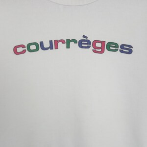 May include: A white ribbed knit top featuring the word courr&egrave;ges printed in an arched design across the chest. The letters alternate between blue, green, and pink colors, creating a vibrant graphic element against the plain white fabric background.