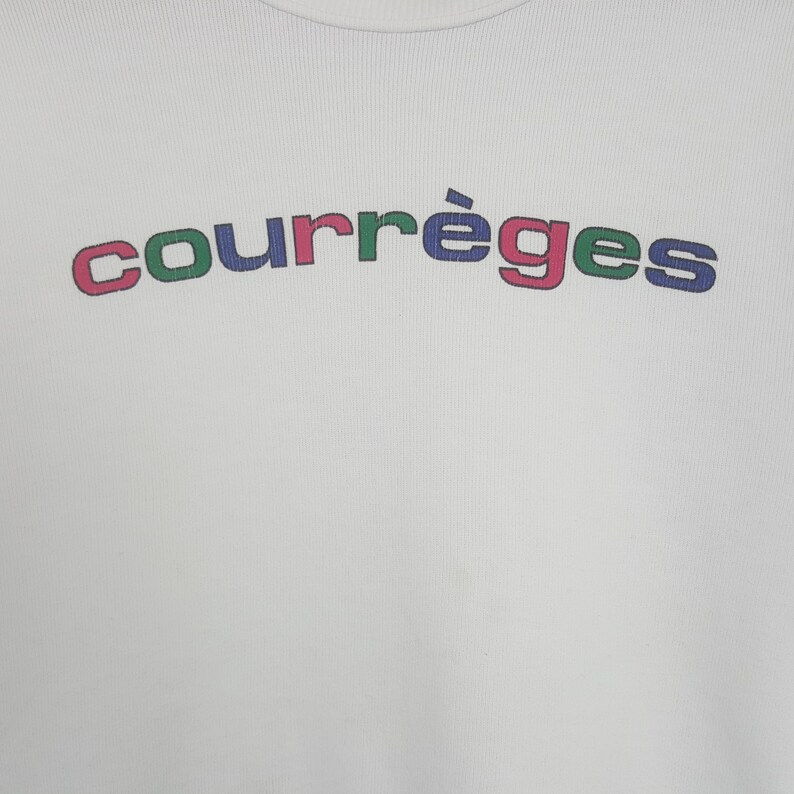 May include: A white ribbed knit top featuring the word courr&egrave;ges printed in an arched design across the chest. The letters alternate between blue, green, and pink colors, creating a vibrant graphic element against the plain white fabric background.
