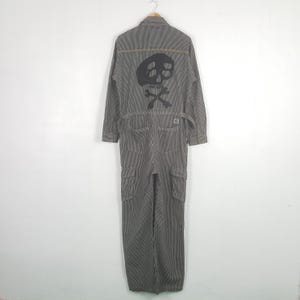May include: A gray and white pinstriped jumpsuit featuring a large black skull and crossbones graphic on the back. The garment includes long sleeves, a collar, two back pockets, and two side cargo pockets on the legs with adjustable waist tabs.