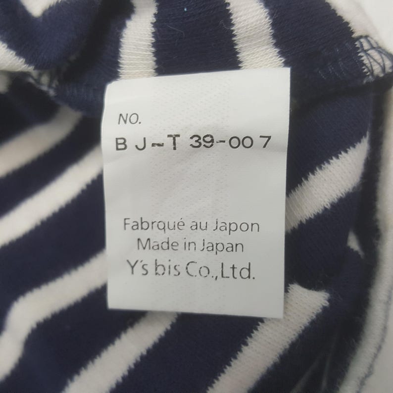 May include: Close-up of a white tag with black text, attached to a navy blue and white striped fabric. The tag reads "NO. BJ-T 39-007", "Fabriqu&eacute; au Japon", "Made in Japan", and "Y's bis Co., Ltd."