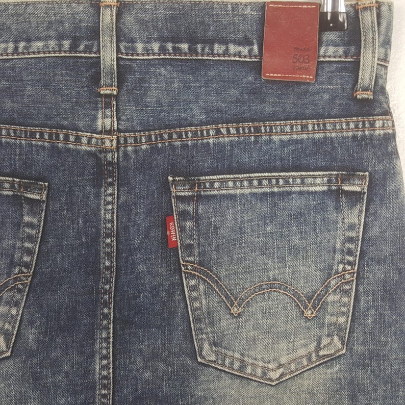Puede incluir: Primer plano de la parte trasera de unos vaqueros azules. Los vaqueros tienen un lavado descolorido y costuras visibles. Una peque&ntilde;a etiqueta roja con la marca "EDWIN" est&aacute; en el bolsillo trasero. Una etiqueta de cuero marr&oacute;n con "Model 503" es visible.